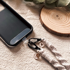May include: Black phone case with a braided gray phone strap. The strap has black metal clasps and small charms with heart and letter designs. A wooden coaster and greenery are in the background.