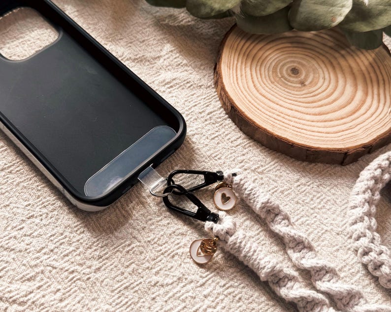 May include: Black phone case with a braided gray phone strap. The strap has black metal clasps and small charms with heart and letter designs. A wooden coaster and greenery are in the background.