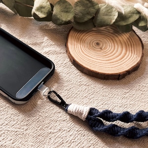 May include: Black phone case with a silver metal attachment and a navy blue and white macrame phone strap. The strap has wooden beads and a black clasp. A wooden slice and greenery are in the background.