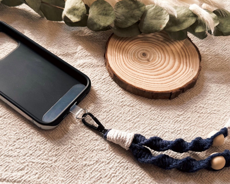 May include: Black phone case with a silver metal attachment and a navy blue and white macrame phone strap. The strap has wooden beads and a black clasp. A wooden slice and greenery are in the background.