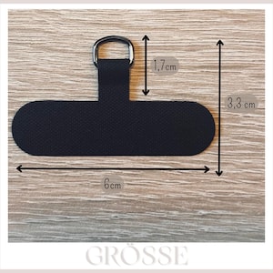 May include: Black fabric phone accessory with a metal ring. The accessory is 6 cm long and 3.3 cm wide, with a ring section that is 1.7 cm long. The accessory is designed to attach to a phone case.