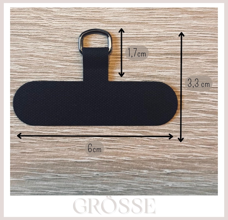 May include: Black fabric phone accessory with a metal ring. The accessory is 6 cm long and 3.3 cm wide, with a ring section that is 1.7 cm long. The accessory is designed to attach to a phone case.