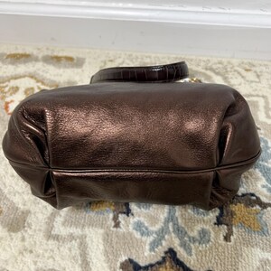 May include: A metallic bronze handbag with a rounded shape and a short handle. The bag has a textured, shiny finish and a dark brown handle with a reptile-like pattern. The bag is sitting on a patterned rug.