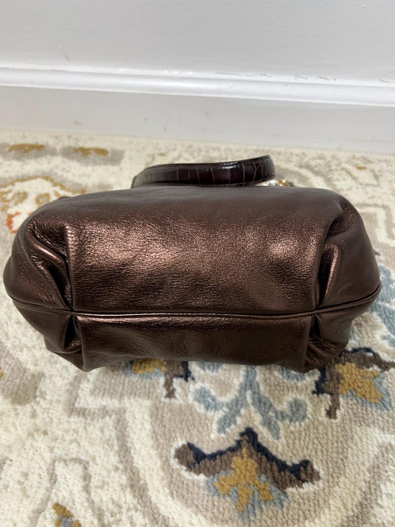 May include: A metallic bronze handbag with a rounded shape and a short handle. The bag has a textured, shiny finish and a dark brown handle with a reptile-like pattern. The bag is sitting on a patterned rug.