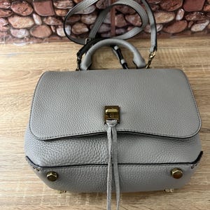 Pu&ograve; includere: Una borsa a mano in pelle grigia con manico superiore e tracolla. La borsa ha una chiusura a patta con chiusura color oro e borchie decorative. La borsa ha una superficie strutturata e un piccolo dettaglio a nappa.