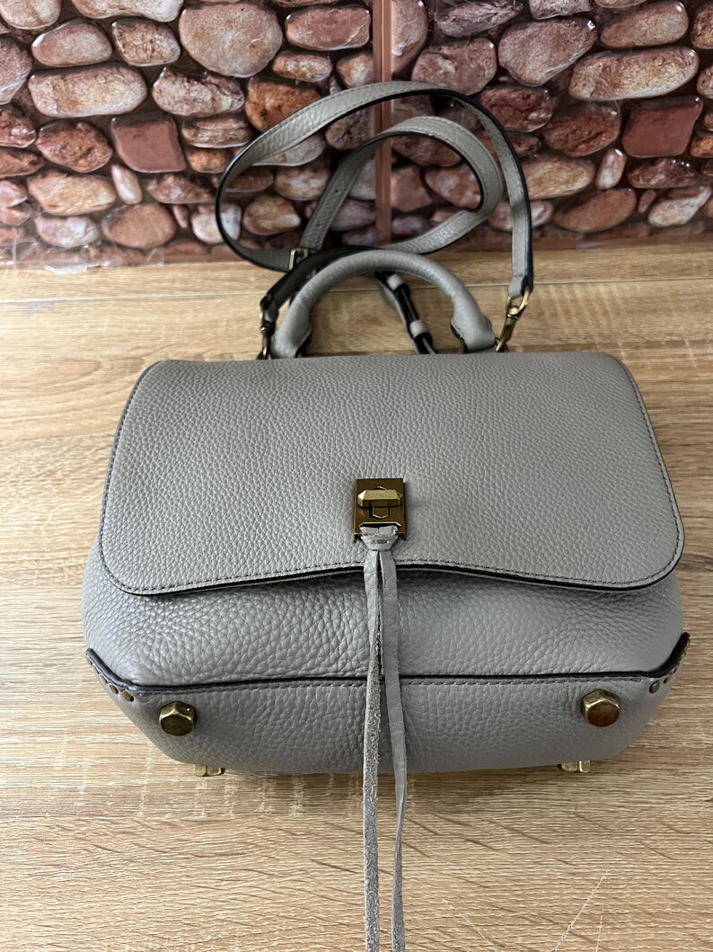 Pu&ograve; includere: Una borsa a mano in pelle grigia con manico superiore e tracolla. La borsa ha una chiusura a patta con chiusura color oro e borchie decorative. La borsa ha una superficie strutturata e un piccolo dettaglio a nappa.