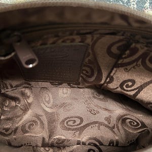 May include: Interior view of a brown handbag with a zipper closure. The inside lining features a patterned design in shades of brown. A small leather tag is visible inside the bag.