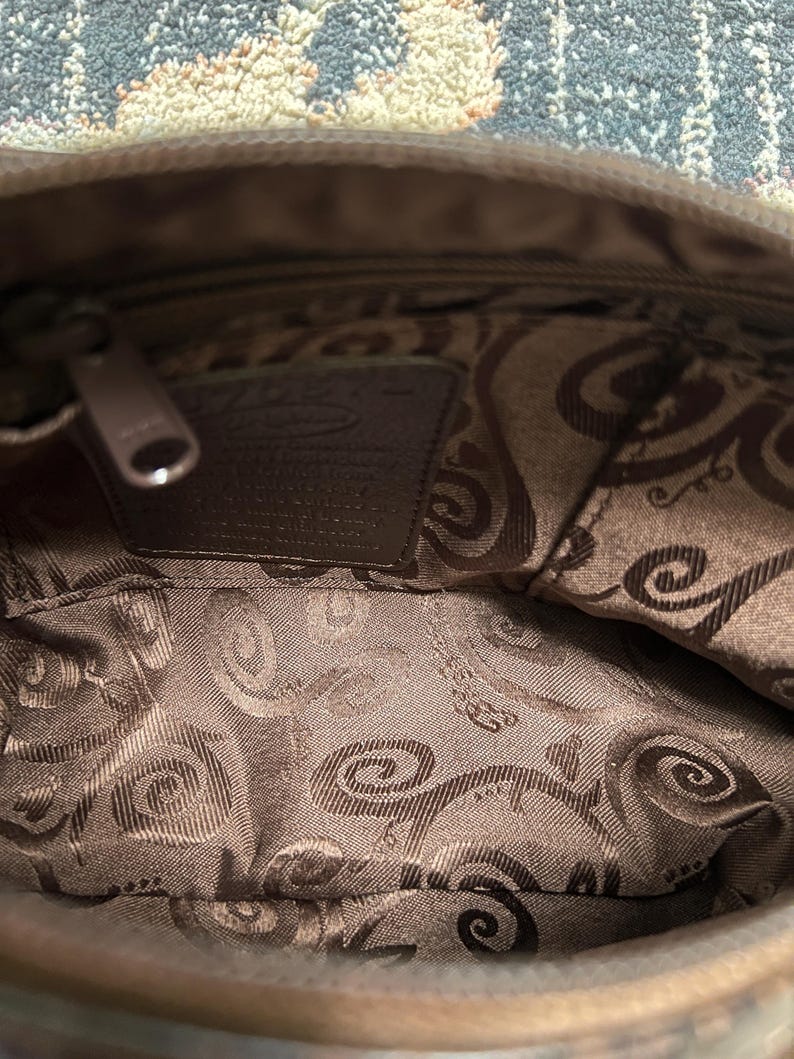 May include: Interior view of a brown handbag with a zipper closure. The inside lining features a patterned design in shades of brown. A small leather tag is visible inside the bag.