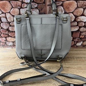 Pu&ograve; includere: Una borsa in pelle grigia con un manico superiore e una tracolla staccabile. La borsa ha una forma rettangolare con una tasca frontale e hardware color oro. La borsa &egrave; appoggiata su una superficie di legno.