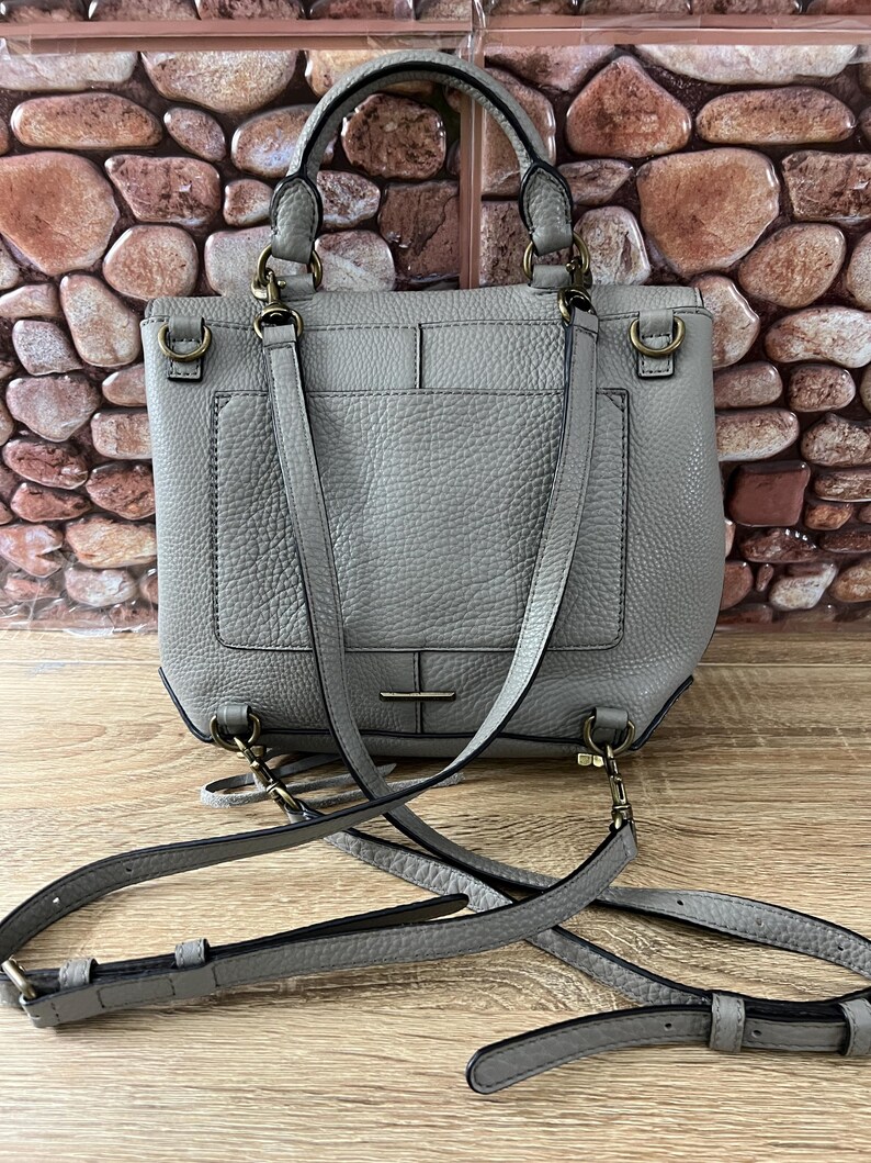 Pu&ograve; includere: Una borsa in pelle grigia con un manico superiore e una tracolla staccabile. La borsa ha una forma rettangolare con una tasca frontale e hardware color oro. La borsa &egrave; appoggiata su una superficie di legno.