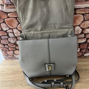 Pu&ograve; includere: Borsa a tracolla in pelle grigia con chiusura a patta e tracolla regolabile. La borsa presenta una finitura strutturata e una chiusura color oro. L'interno &egrave; foderato con un tessuto grigio pi&ugrave; chiaro. La borsa &egrave; aperta.