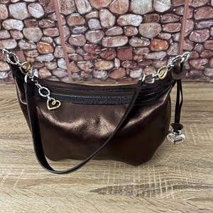 May include: A metallic brown shoulder bag with a curved shape. The bag has a black zipper and a long, thin strap. Silver-toned chain and heart-shaped charms adorn the bag. The background features a stone wall.