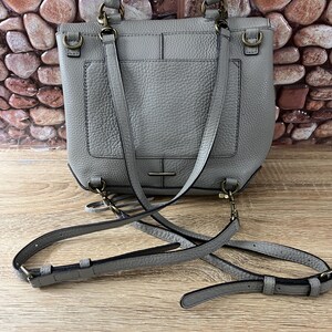 Pu&ograve; includere: Una borsa in pelle grigia con manico superiore e tracolla staccabile. La borsa ha una forma rettangolare con una tasca frontale e hardware color oro. La borsa &egrave; appoggiata su una superficie di legno.