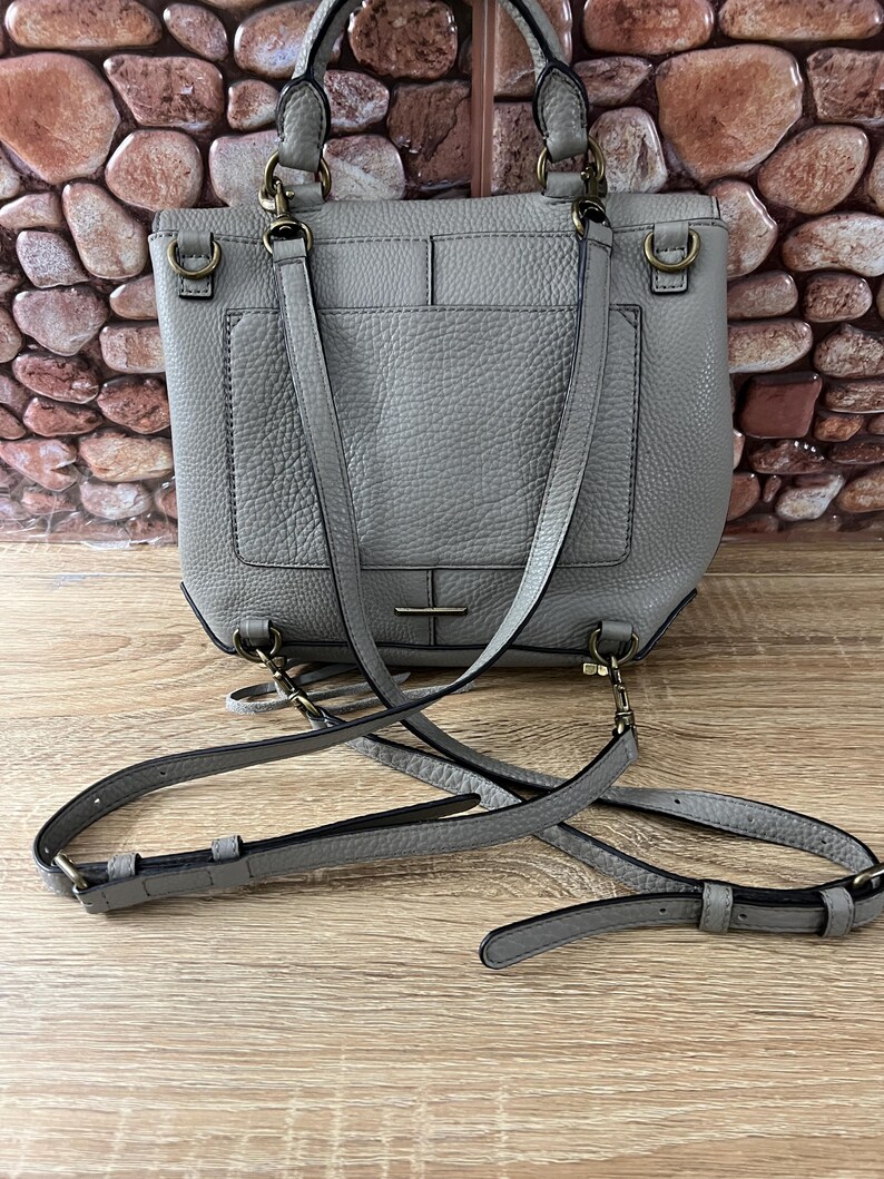 Pu&ograve; includere: Una borsa in pelle grigia con manico superiore e tracolla staccabile. La borsa ha una forma rettangolare con una tasca frontale e hardware color oro. La borsa &egrave; appoggiata su una superficie di legno.