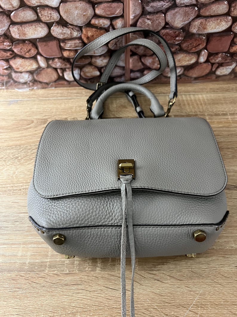 Pu&ograve; includere: Una borsa a mano in pelle grigia con manico superiore e tracolla. La borsa ha una chiusura a patta con una chiusura color oro e borchie decorative. La borsa ha una superficie strutturata e un piccolo dettaglio a nappa.