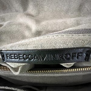 Pu&ograve; includere: Primo piano dell'interno di una borsa in pelle grigia. L'etichetta in pelle nera mostra il nome del marchio "REBECCA MINKOFF" in lettere in rilievo. &Egrave; visibile una cerniera dorata e l'interno della borsa &egrave; in un materiale grigio chiaro e morbido.