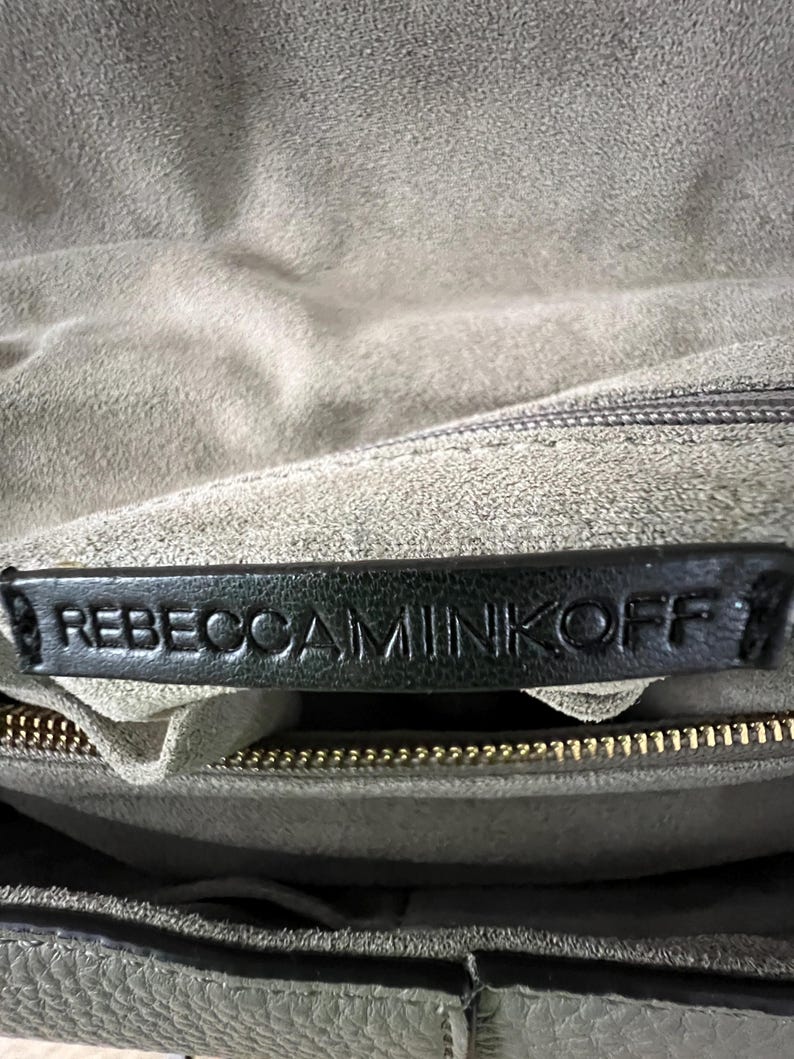 Pu&ograve; includere: Primo piano dell'interno di una borsa in pelle grigia. L'etichetta in pelle nera mostra il nome del marchio "REBECCA MINKOFF" in lettere in rilievo. &Egrave; visibile una cerniera dorata e l'interno della borsa &egrave; in un materiale grigio chiaro e morbido.