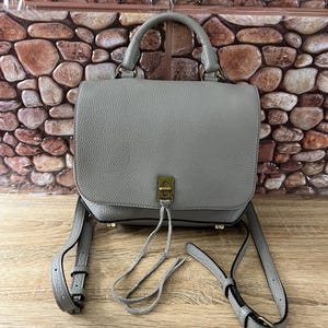 Pu&ograve; includere: Una borsa a mano in pelle grigia con un manico superiore e una lunga tracolla regolabile. La borsa ha una chiusura a patta con un fermaglio color oro e un nappa decorativa. La borsa &egrave; su una superficie di legno.