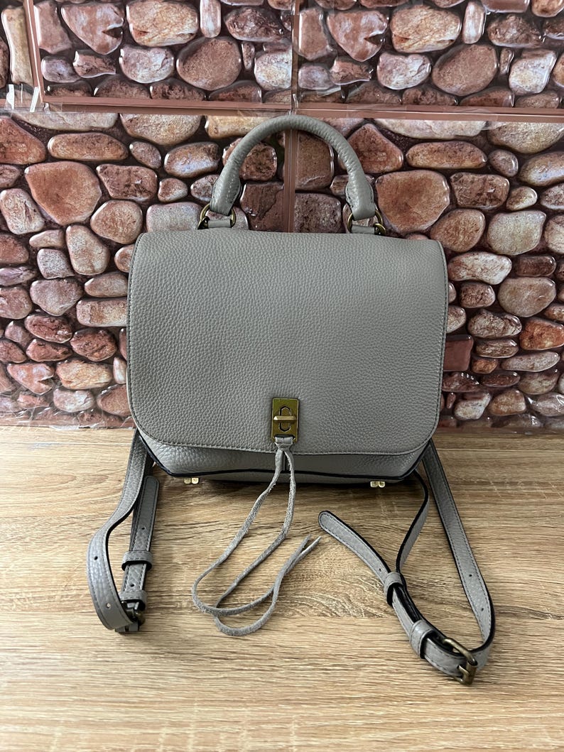 Pu&ograve; includere: Una borsa a mano in pelle grigia con un manico superiore e una lunga tracolla regolabile. La borsa ha una chiusura a patta con un fermaglio color oro e un nappa decorativa. La borsa &egrave; su una superficie di legno.