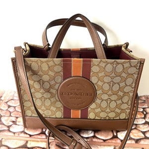 May include: A brown and tan tote bag featuring the Coach signature monogram pattern, a central vertical orange and maroon stripe, and a circular leather patch embossed with the text Coach New York. It includes two top handles and a shoulder strap.
