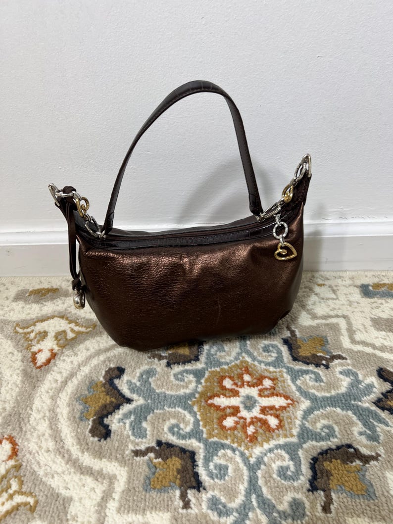 May include: A metallic brown handbag with a curved handle and silver-toned hardware. The bag has a zipper closure and a small charm. The bag is sitting on a patterned rug.