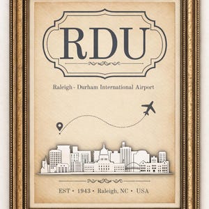 Könnte beinhalten: Gerahmter Druck mit den Buchstaben "RDU", die den Raleigh-Durham International Airport darstellen. Das Bild zeigt eine Skyline, ein Flugzeug und den Text "EST 1943 Raleigh, NC USA". Der Rahmen ist goldfarben.