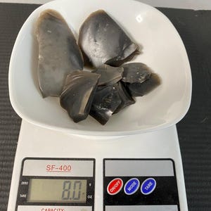 May include: A white digital scale with a black display showing 8.0 ounces. The scale is on a black surface and has a white bowl with several pieces of gray flint in it.