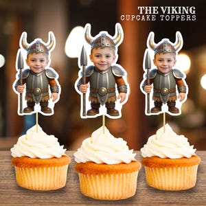 Op de afbeelding: Drie cupcakes met witte glazuur, elk bekroond met een Viking-thema cupcake topper. De toppers tonen een cartoonafbeelding van een kind verkleed als Viking, met een speer. De tekst "THE VIKING CUPCAKE TOPPERS" staat bovenaan.