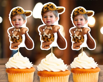 May include: Three cupcakes with white frosting, each topped with a cartoon cutout of a baby dressed as a Flintstones character. The baby wears a leopard-print hat and loincloth, holding a wooden club. Text at the top reads "Bamm Bamm-Flintstones Cupcake Toppers."