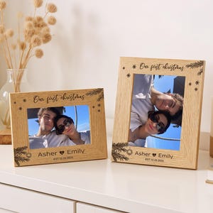 May include: Two light-colored wooden picture frames with horizontal and vertical orientations. Each frame is engraved with the text Our first christmas, the names Asher and Emily, and the date EST.12.25.2025, featuring pine branch and snowflake motifs.