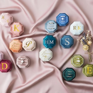 May include: A collection of velvet ring boxes in shades like pink, blue, and green, displayed on a pink satin cloth. Some boxes have gold initials or names, while others contain rings. A small bunch of dried flowers is also visible in the image.