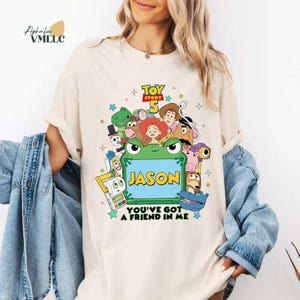 May include: Cream-coloured t-shirt with a vibrant graphic of Toy Story 5 characters. The design includes a green frog with the name "JASON" and the phrase "YOU'VE GOT A FRIEND IN ME". The shirt has short sleeves and a crew neck.