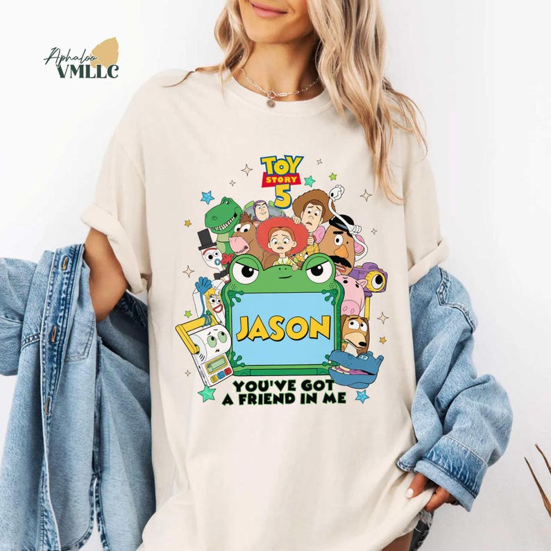 May include: Cream-coloured t-shirt with a vibrant graphic of Toy Story 5 characters. The design includes a green frog with the name "JASON" and the phrase "YOU'VE GOT A FRIEND IN ME". The shirt has short sleeves and a crew neck.