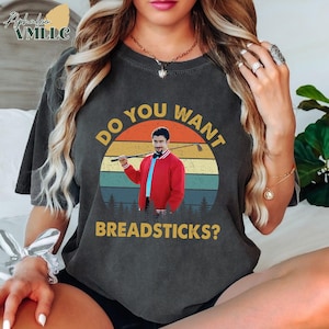 May include: Dark gray t-shirt with a retro graphic of a man in a red jacket holding a golf club, set against a sunset backdrop. The text "DO YOU WANT BREADSTICKS?" is arched above and below the image. The shirt has a relaxed fit.