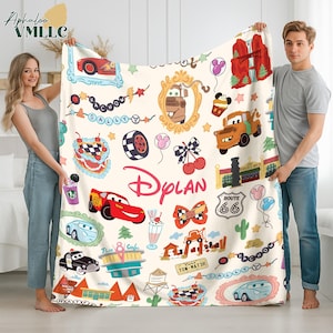 A man and a woman are holding up a large blanket with various cartoon car designs and other car-related items, such as a fire hydrant, traffic light, and gas station.