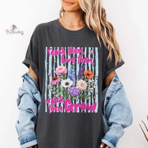 May include: Dark gray t-shirt with a floral design and the text "Teach them, love them, watch them Grow." The design features a bouquet of colorful flowers against a striped background.
