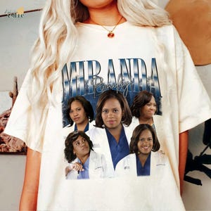 May include: Cream-colored t-shirt featuring a graphic of Miranda Bailey with the name "Miranda Bailey" in a blue and white design. The graphic includes multiple images of the character. The shirt is a casual style.