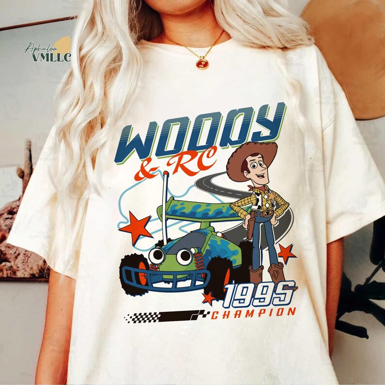 Op de afbeelding: Cr&egrave;mekleurig T-shirt met een afbeelding van Woody en een RC-auto met de tekst "WOODY & RC 1995 CHAMPION". Het ontwerp bevat een cartoonafbeelding van Woody met een cowboyhoed en een blauw-groene RC-auto.