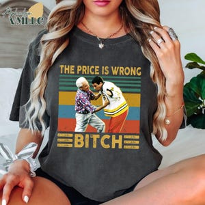 May include: Dark gray t-shirt with a graphic featuring a retro design. The design includes the text "THE PRICE IS WRONG BITCH" above an image of two men in a confrontation. The shirt has a vintage, worn look.