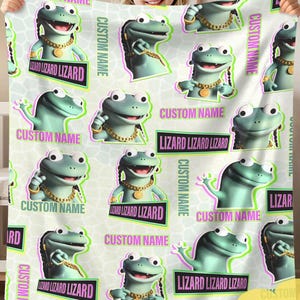 May include: A white blanket featuring a repeating pattern of cartoon lizards with gold chains and braids. The lizards are light green with black outlines. Text includes "CUSTOM NAME" and "LIZARD LIZARD LIZARD".