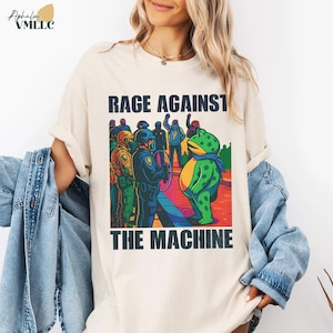 May include: Cream-colored t-shirt featuring the text "RAGE AGAINST THE MACHINE" above a colorful graphic. The graphic depicts a frog facing a line of police officers. The frog is green with a blue scarf and yellow belly.
