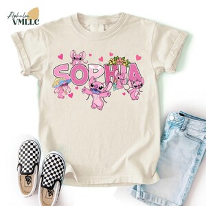 A beige t-shirt with a personalized name printed on it, featuring cartoon characters and hearts. The t-shirt is placed next to a pair of checkered shoes and a pair of denim shorts.