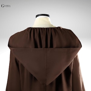 Pu&ograve; includere: Un mantello con cappuccio marrone scuro caratterizzato da uno scollo arricciato e un ampio cappuccio a punta. Il capo &egrave; esposto su un manichino, con il testo GLORIA visibile nell'angolo in alto a sinistra. Il tessuto ha una consistenza opaca.
