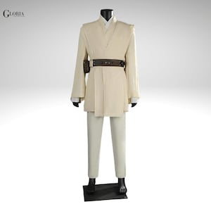 Pu&ograve; includere: Un completo composto da tunica e pantaloni color crema, caratterizzato da un top a portafoglio con maniche lunghe, una cintura utility marrone scuro con una piccola tasca attaccata e una maglietta bianca. La parola GLORIA &egrave; stampata in nero in alto a sinistra.