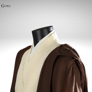 Pu&ograve; includere: Un indumento a strati composto da una tunica interna color crema con scollo a V e una tunica esterna marrone scuro con maniche larghe. Il testo GLORIA CUSTOM MADE &egrave; visibile nell'angolo in alto a sinistra su un manichino nero.