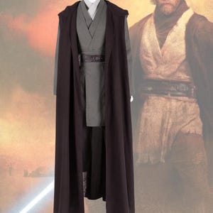 May include: A gray costume with a long, dark brown hooded cloak. The costume includes a gray tunic, pants, and a belt. The costume is displayed on a mannequin. The background features a blurred image of a person.