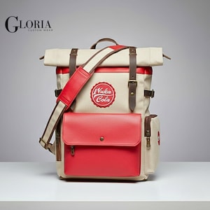 Pode incluir: Uma mochila bege e vermelha com detalhes em couro castanho. A mochila apresenta um bolso frontal vermelho e uma alça de ombro. O logotipo "Nuka Cola" é exibido na frente. A bolsa tem uma alça superior e alças ajustáveis.