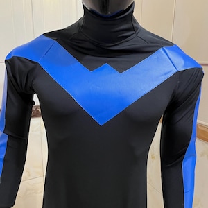 Puede incluir: Disfraz de superhéroe negro y azul con cuello alto. El disfraz presenta detalles azules en los hombros y los brazos, y un diseño de cheurón en el pecho. El atuendo de manga larga se exhibe en un maniquí.