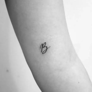 Op de afbeelding: Een kleine tatoeage van de cursieve letter "B" in zwarte inkt. De tatoeage staat op een lichte huidskleur, waarbij de elegante scriptstijl van de letter duidelijk zichtbaar is. Het ontwerp is eenvoudig en minimalistisch.