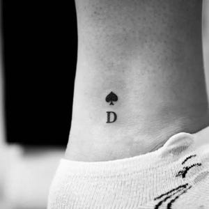 Op de afbeelding: Een kleine, zwarte inkt-tatoeage op de huid met het schoppensymbool van een speelkaart direct boven de hoofdletter D. Het ontwerp is uitgevoerd in een eenvoudig, strak lettertype en een effen zwarte vulling voor een helder visueel beeld.